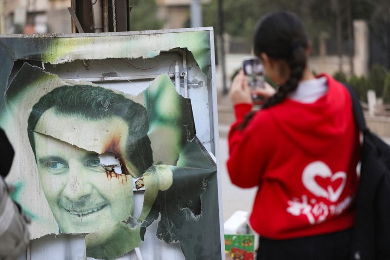 © Reuters. A woman uses her mobile phone near a damaged picture of Syrian President Bashar al-Assad as people celebrate, after Syrian rebels announced that they have ousted President Bashar al-Assad, in Qamishli, Syria December 8, 2024. REUTERS/Orhan Qereman